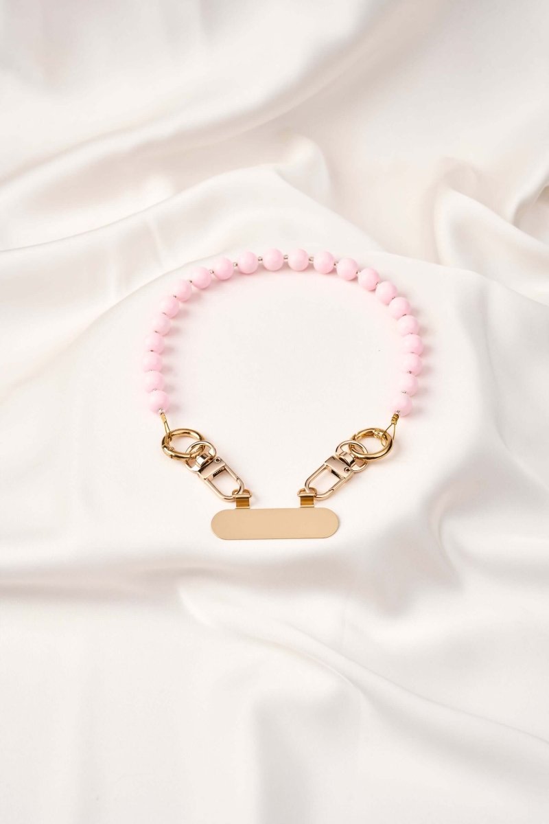 strap celular artesanal Blush Moment en tonos rosa con broches dorados sobre tela blanca.
