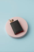 Mini tarjetero negro artesanal sobre una base circular rosa, con broche dorado y material de PVC.