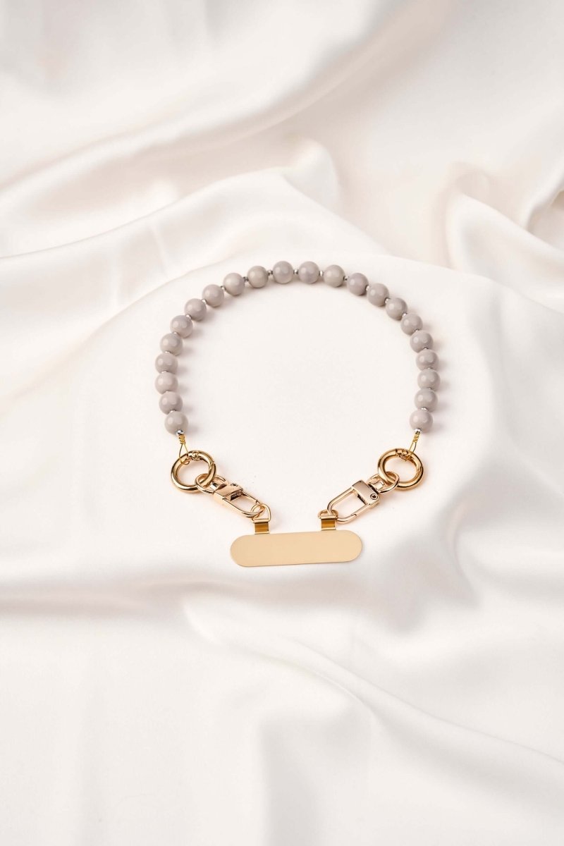 strap celular artesanal Silver Calm en tonos grises con broches dorados sobre tela blanca.